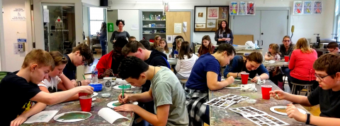 An art studio where two adults are overseeing tables of children painting.