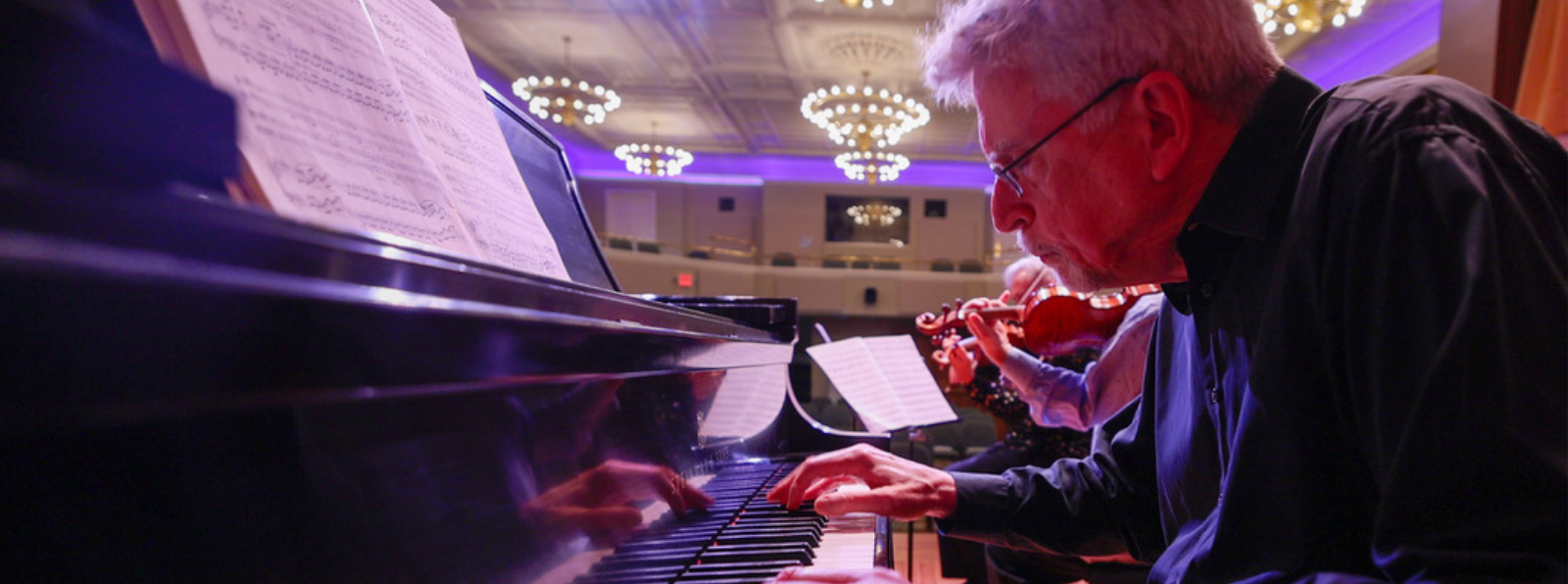 A pianist in a concert hall, accompanied by other instrumentalists behind him.