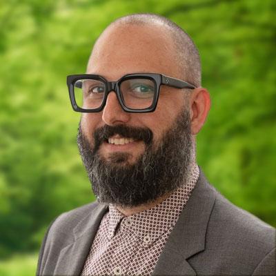 Kirk Amaral Snow headshot. Man smiling with glasses, beard, a dark grey sport coat and brown shirt.  