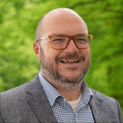 Ryan Patterson headshot. Smiling man with glasses, a mustache and beard, and wearing a grey sport coat over a light blue shirt.