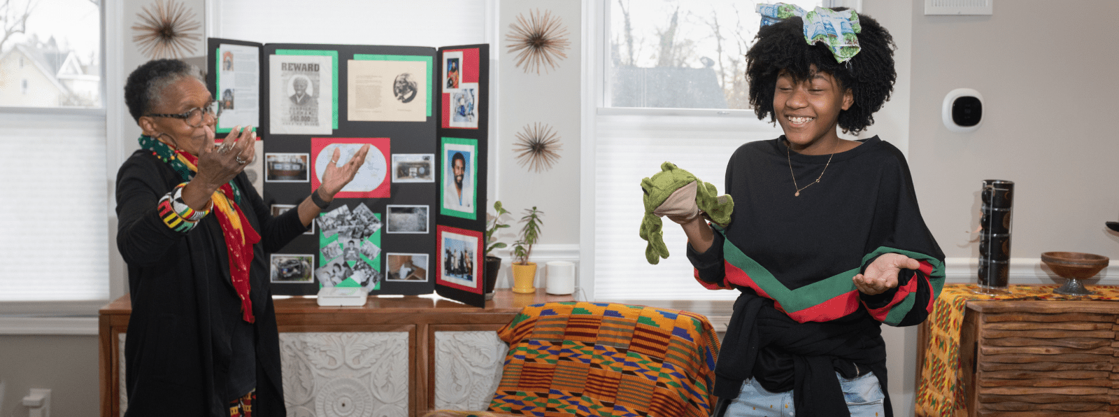 An adult raises their hands to a child with a frog hand puppet.